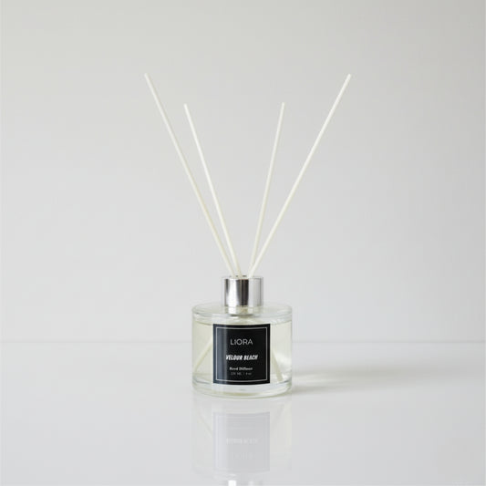 Liora Scentique Velour Beach reed diffuser with white reeds on driftwood, shells, and lavender, with the ocean and beach in the background.
