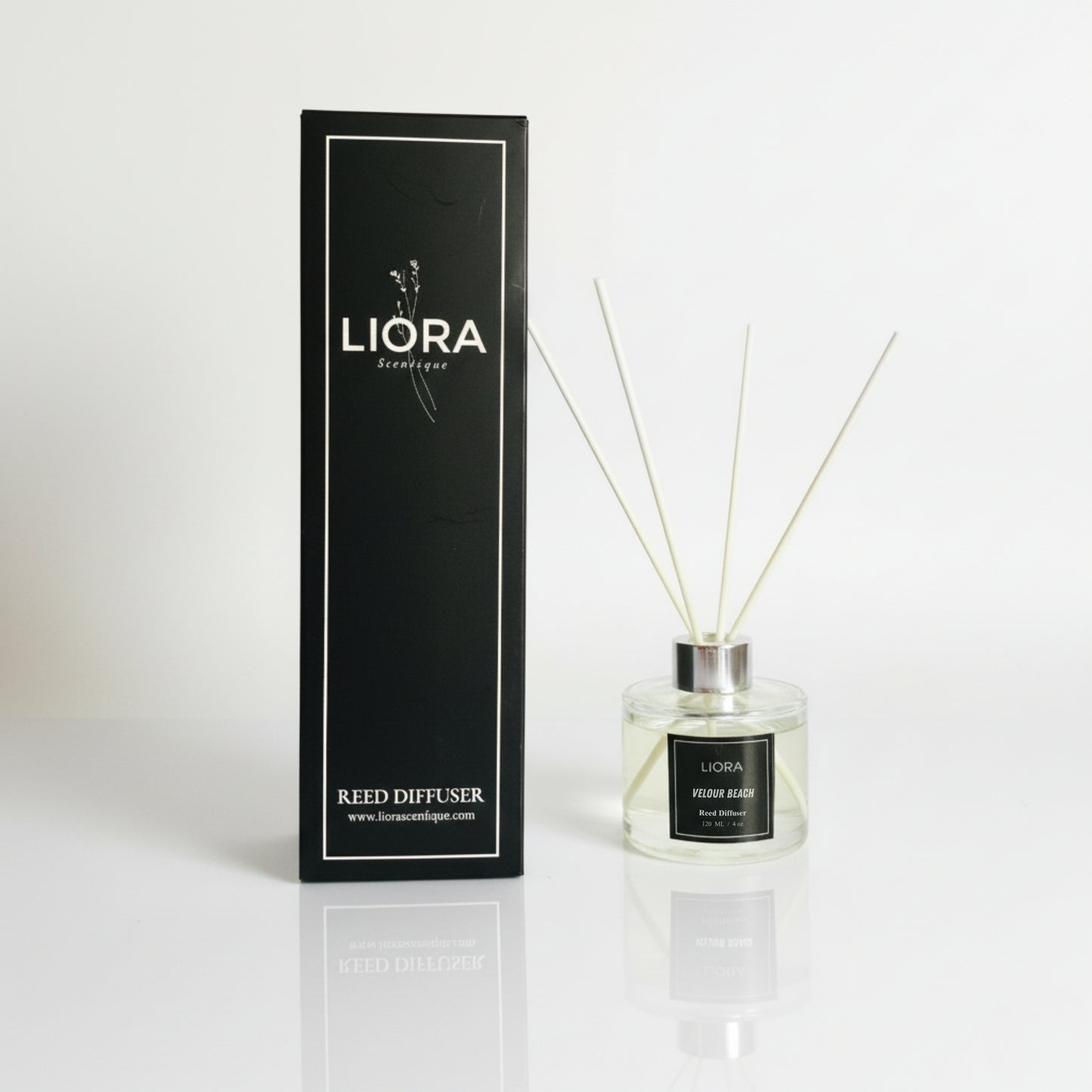 Liora Scentique Velour Beach reed diffuser with white reeds on driftwood, shells, and lavender, with the ocean and beach in the background.