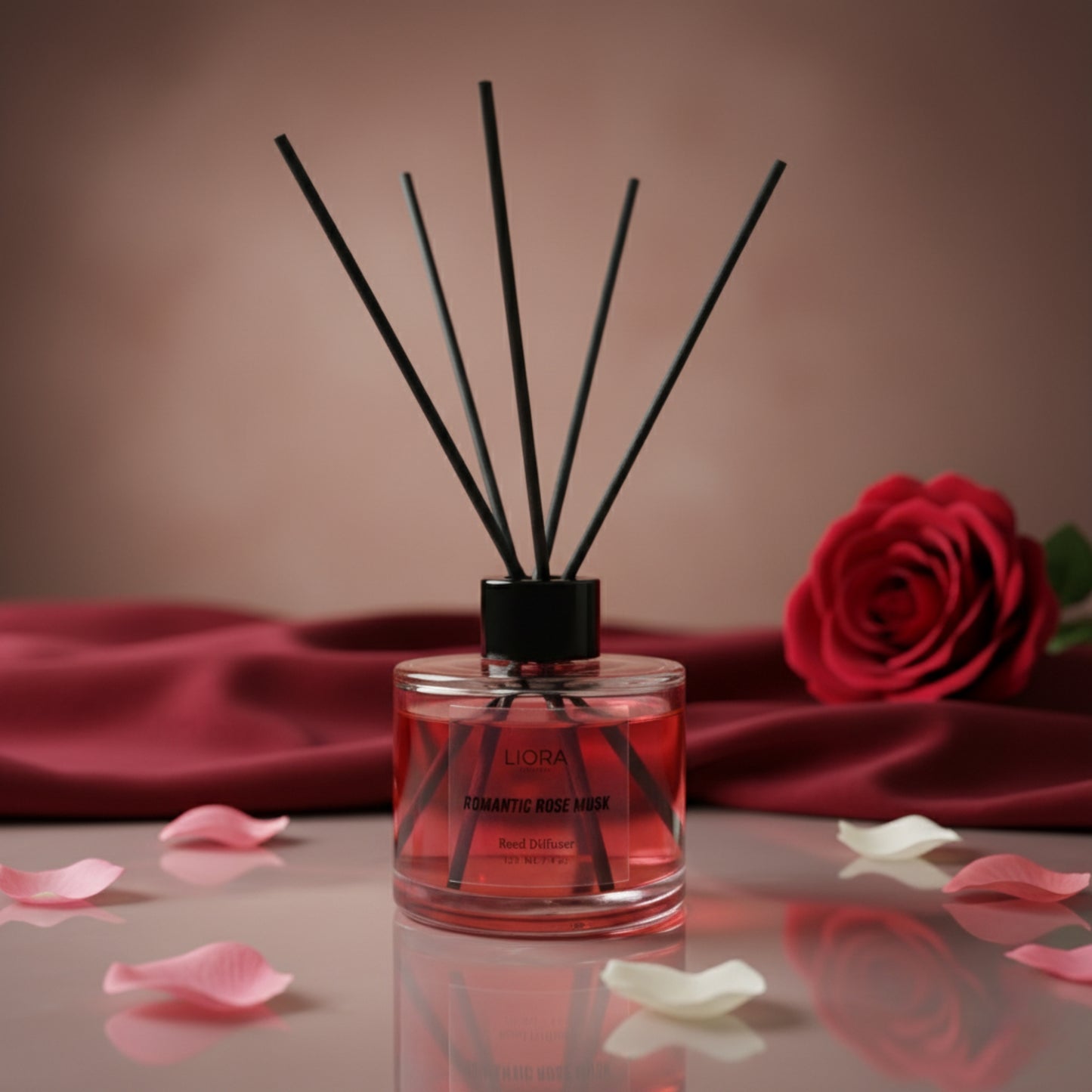 Reed diffuser with black sticks and pink liquid on a reflective surface with a red rose and petals in the background.