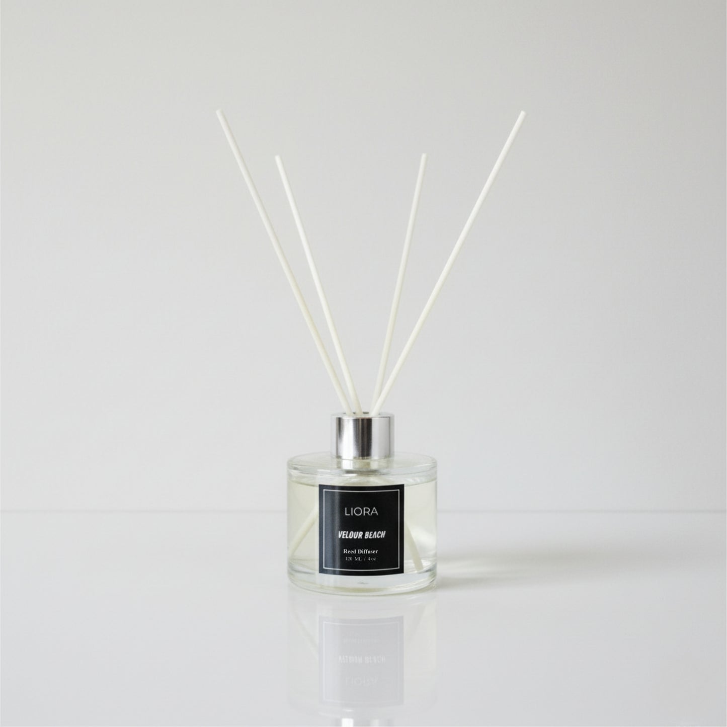 Liora Scentique Velour Beach reed diffuser with white reeds on driftwood, shells, and lavender, with the ocean and beach in the background.