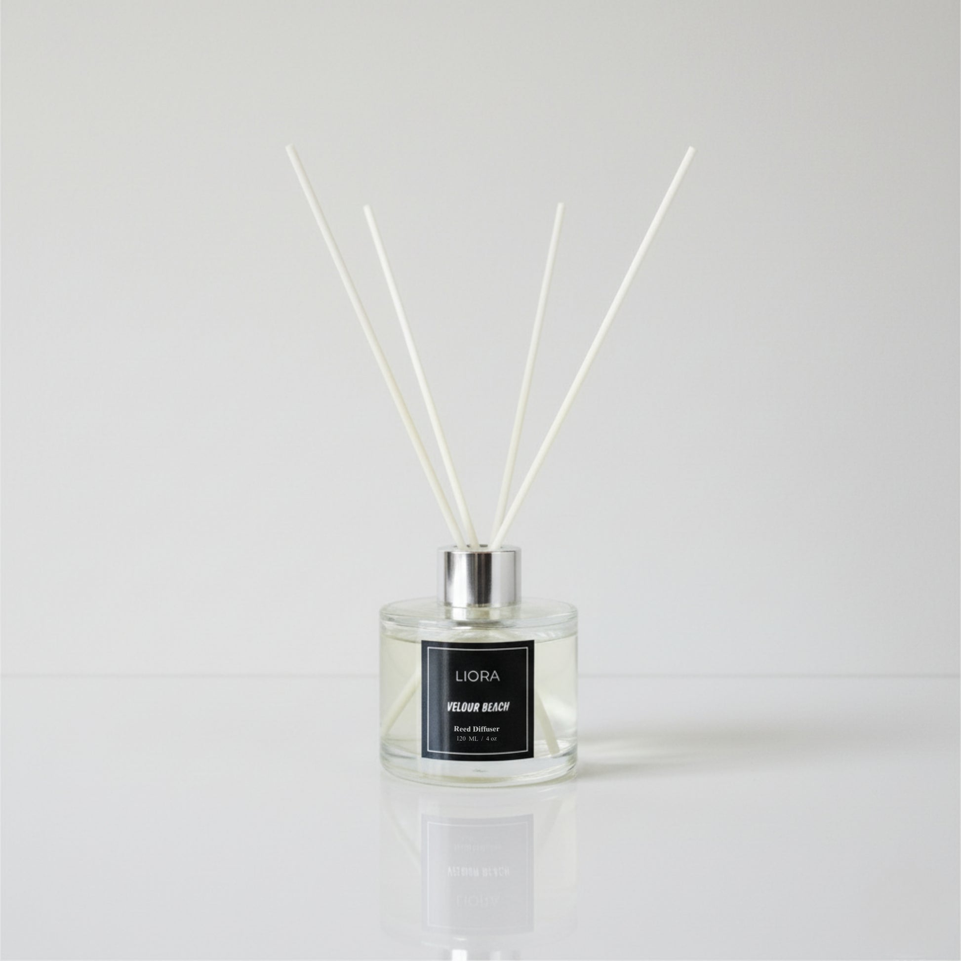 Liora Scentique Velour Beach reed diffuser with white reeds on driftwood, shells, and lavender, with the ocean and beach in the background.