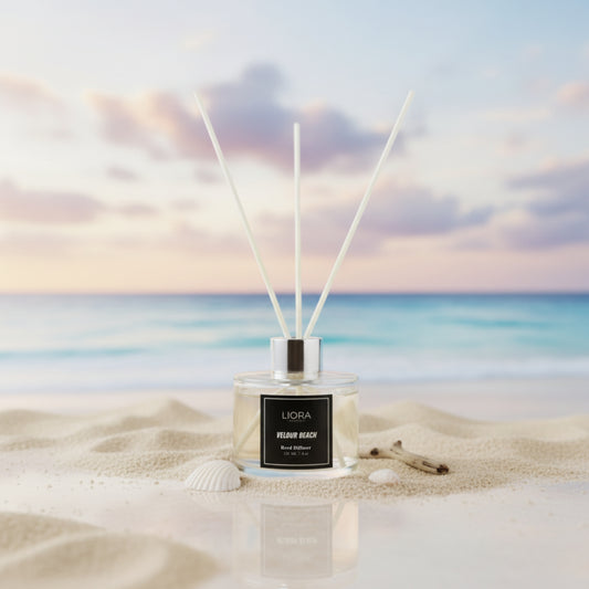 Liora Scentique Velour Beach reed diffuser with white reeds on driftwood, shells, and lavender, with the ocean and beach in the background.